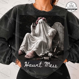 May include: A charcoal gray mineral-washed sweatshirt featuring a graphic of a ghost wearing pink hair rollers and fuzzy slippers, holding a wine glass while sitting by a tombstone with wine bottles. The text Haunt Mess is printed below.