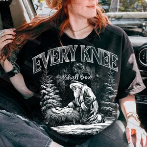 May include: Black t-shirt with a graphic design featuring the text "EVERY KNEE Shall Bow". The design includes an illustration of Santa Claus kneeling before a manger, with pine trees on either side.