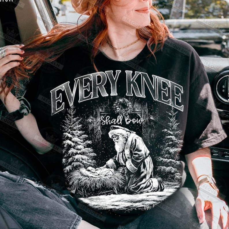 May include: Black t-shirt with a graphic design featuring the text "EVERY KNEE Shall Bow". The design includes an illustration of Santa Claus kneeling before a manger, with pine trees on either side.