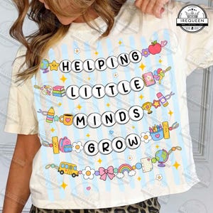 May include: A cream-colored t-shirt featuring a light blue vertical striped pattern and a graphic print with the text HELPING LITTLE MINDS GROW. The design includes colorful illustrations of school supplies, a globe, a bus, and a rainbow.
