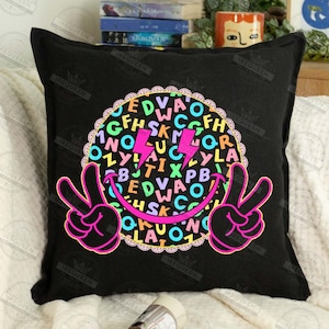 May include: Black square pillow with a colorful graphic. The design features a smiley face with peace signs, a circle filled with letters, and two pink lightning bolts. The pillow is on a white surface.