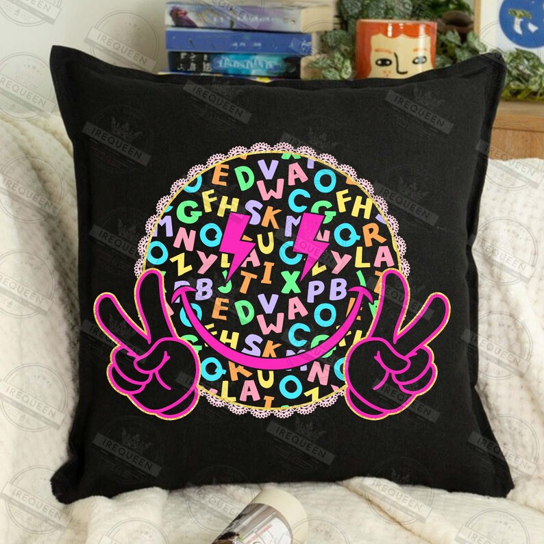 May include: Black square pillow with a colorful graphic. The design features a smiley face with peace signs, a circle filled with letters, and two pink lightning bolts. The pillow is on a white surface.