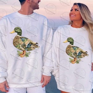 May include: White sweatshirts featuring a detailed duck illustration. The duck has a green head, yellow beak, and a body with a patterned design. The sweatshirts are long-sleeved and suitable for casual wear.