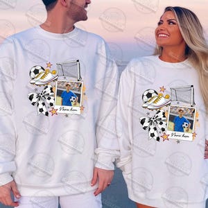 Puede incluir: Sudadera blanca con un dise&ntilde;o de tem&aacute;tica futbol&iacute;stica. El dise&ntilde;o presenta un bal&oacute;n de f&uacute;tbol, botas, una porter&iacute;a y un marco de fotos con un jugador de f&uacute;tbol. El texto "Name here" est&aacute; impreso debajo del marco de fotos.