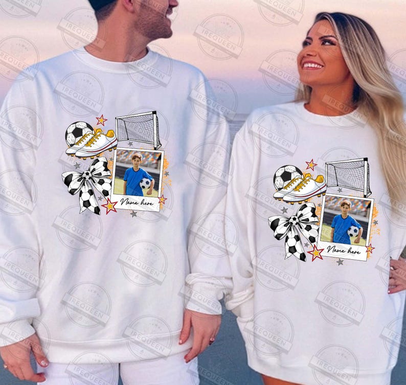 Puede incluir: Sudadera blanca con un dise&ntilde;o de tem&aacute;tica futbol&iacute;stica. El dise&ntilde;o presenta un bal&oacute;n de f&uacute;tbol, botas, una porter&iacute;a y un marco de fotos con un jugador de f&uacute;tbol. El texto "Name here" est&aacute; impreso debajo del marco de fotos.