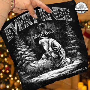 May include: Black graphic tee featuring a detailed illustration of Santa Claus kneeling before the baby Jesus in a manger. The text "EVERY KNEE Shall Bow" is at the top, with "Philippians 2:10" at the bottom. The design is set in a snowy forest scene.