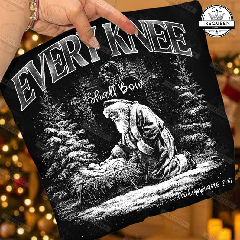 May include: Black graphic tee featuring a detailed illustration of Santa Claus kneeling before the baby Jesus in a manger. The text "EVERY KNEE Shall Bow" is at the top, with "Philippians 2:10" at the bottom. The design is set in a snowy forest scene.