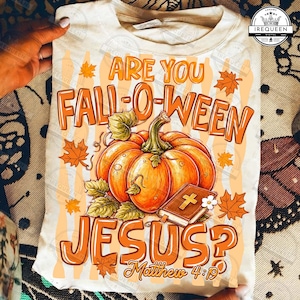 May include: A cream-colored t-shirt featuring a graphic print with the orange text ARE YOU FALL-O-WEEN JESUS? and the scripture reference Matthew 4:19. The design includes a large orange pumpkin, green leaves, and a brown book with a gold cross.
