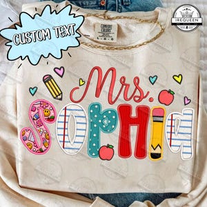 Può includere: Una felpa girocollo color crema con la scritta "Mrs. Sophia" in un carattere multicolore con vari motivi. Il design include grafiche a tema scolastico come una matita, mele e piccoli cuori, con un'etichetta del marchio visibile.
