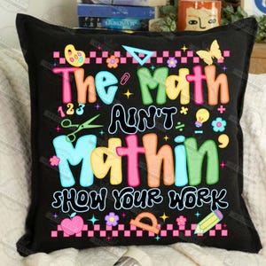 Puede incluir: Coj&iacute;n cuadrado negro con el texto "The Math Ain't Mathin' Show Your Work" en letras coloridas. El dise&ntilde;o incluye s&iacute;mbolos matem&aacute;ticos, una mariposa, flores y un l&aacute;piz.