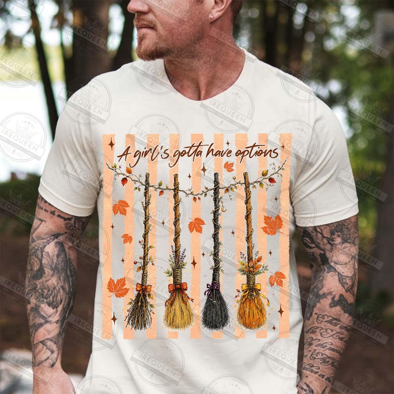 May include: A cream-colored short-sleeve t-shirt featuring a graphic print of four distinct broomsticks hanging from a string. The text "A girl's gotta have options" is printed above the brooms, surrounded by orange maple leaves and star accents.