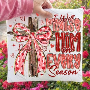 May include: White square pillow with a cross design. The cross is made of wood and has a pink bow with red hearts. The text "I Will Praise Him in Every Season" is in red. The text "Psalm 146:2" is also visible.
