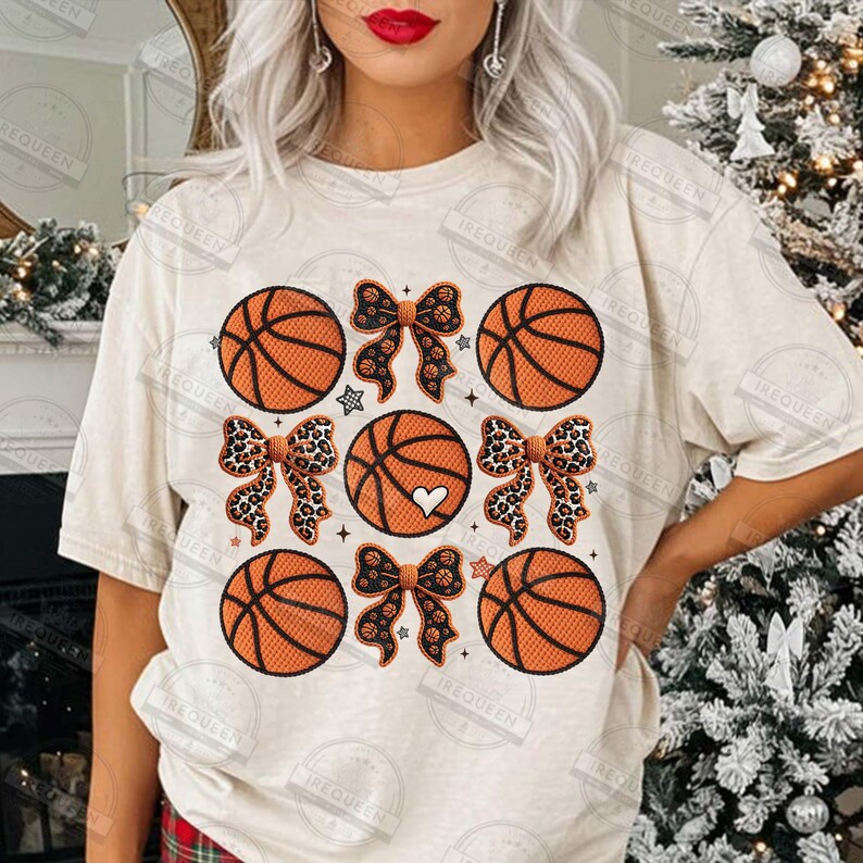 May include: Cream-colored t-shirt featuring a basketball-themed design. The design includes orange basketballs, leopard print bows, and a heart. The shirt has a crew neck and short sleeves. The text "IREQUEEN" is visible on the shirt.