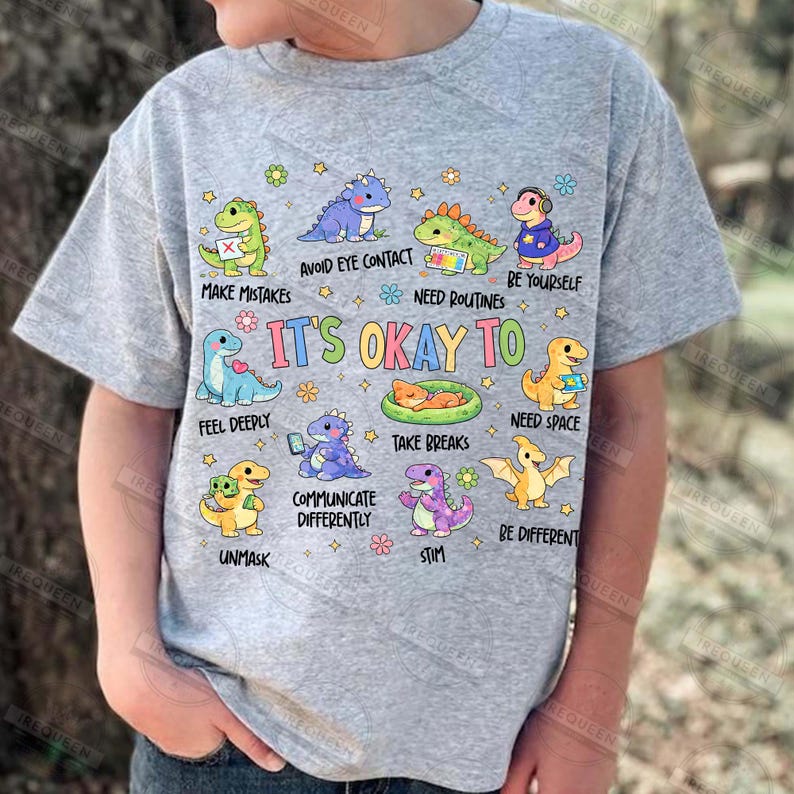 May include: A grey t-shirt with cartoon dinosaurs and phrases such as "Avoid Eye Contact" and "It's Okay To." The shirt has a round neck and short sleeves, suitable for casual wear.