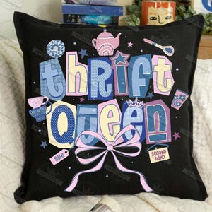May include: Black throw pillow with the words "thrift queen" in colorful, whimsical lettering. The design includes illustrations of a teapot, teacup, stars, and a bow. The pillow has a tag that says "sale".