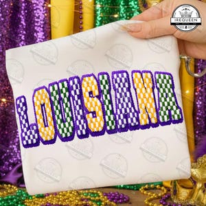 May include: A white fabric item with the embroidered word "LOUISIANA" in purple, yellow, and green checkered letters. The design is set against a backdrop of purple and gold Mardi Gras beads and decorations. The image includes the "IREQUEEN" logo.