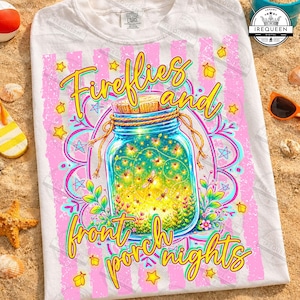 May include: White t-shirt featuring a vibrant graphic of a mason jar filled with glowing fireflies. The design includes the words "Fireflies and front porch nights" in a playful font, surrounded by stars and floral elements.