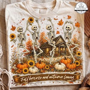May include: A cream-colored t-shirt featuring a graphic of four skeletons dancing in an autumn landscape with pumpkins, sunflowers, and a cabin. A brown banner at the bottom displays the text "Fall breeze and autumn leaves" in white script.