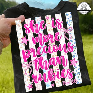 Può includere: T-shirt nera con la scritta rosa "She is more precious than rubies" e la citazione biblica Proverbi 3:15. Il design include strisce bianche e nere, motivi floreali, stelle e fiocchi rosa.