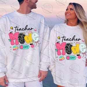 May include: White long-sleeved shirts with a graphic print of the words "Teacher Mode" in pink, yellow, and black with a light blue on/off switch. The graphic includes a variety of school-themed icons such as a lightbulb, apple, pencil, and books.
