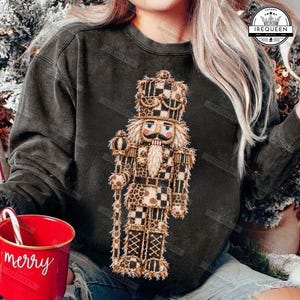 Puede incluir: Una sudadera de cuello redondo gris carbón con un gráfico central de un soldado cascanueces con un patrón de cuadros y leopardo en tonos beige y marrón. Una taza roja con el texto blanco merry es visible en la esquina inferior izquierda.