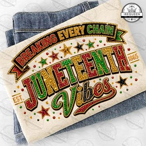 May include: A cream-colored t-shirt with the words "BREAKING EVERY CHAIN" and "JUNETEENTH Vibes" in a decorative font. The design includes stars and the year "1865". The shirt is folded on top of a pair of blue jeans.