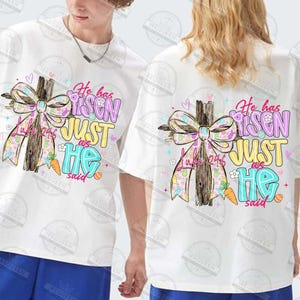 May include: White t-shirts with a colorful graphic featuring a wooden cross, a patterned bow, and the text "He has Risen, Just as He said." The design includes pastel colors and Easter-themed elements.
