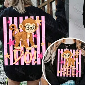 Puede incluir: Sudadera negra con un mono de dibujos animados con gafas, sobre rayas verticales rosas y negras. Las palabras "DO!" y "SEE" est&aacute;n debajo del mono, con acentos de estrellas.