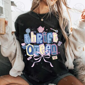 May include: Black sweatshirt with the text "Thrift Queen" in a colorful, whimsical design. The design includes a bow, stars, and illustrations of a teapot, teacup, and other thrift store-related items. The words "sale" and "second hand" are also included.