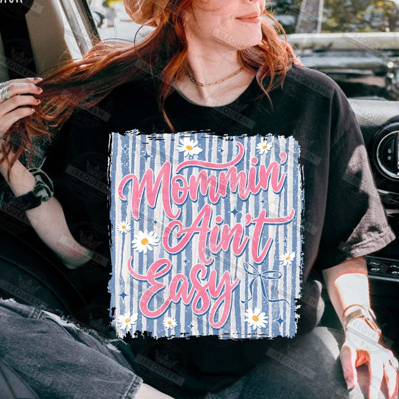 May include: Black t-shirt with a graphic that reads "Mommin' Ain't Easy" in pink script over a blue and white striped background. White daisies and a small bow accent the design. The shirt is a casual style.
