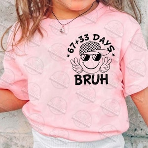 Pu&ograve; includere: Una maglietta rosa chiaro con un design grafico. Il design include una faccina sorridente con occhiali da sole e un cappello a scacchi, con la scritta "67+33 DAYS BRUH". La maglietta &egrave; a maniche corte.