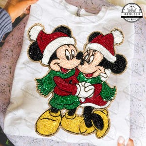 Pode incluir: Camiseta branca de manga comprida com um design de Mickey Mouse e Minnie Mouse com contas. Os personagens usam chapéus de Papai Noel, roupas vermelhas e verdes e dão as mãos. A camisa tem o texto "IREQUEEN".