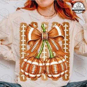 May include: A beige long-sleeve sweatshirt featuring a graphic print of two pumpkins designed to look like brown footballs with white laces. A large brown bow sits atop the pumpkins, and the background includes vertical brown football lace patterns.