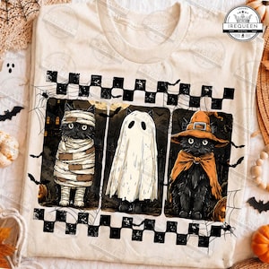 May include: A cream-colored t-shirt featuring a graphic of three black cats in Halloween costumes: a mummy, a ghost, and a witch. The design is framed by a black and white checkered border and includes spiderwebs and bats. The logo IREQUEEN is visible.