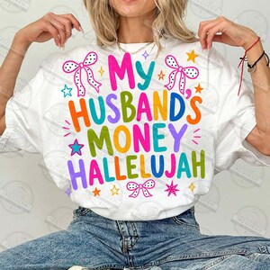 Pu&ograve; includere: Una t-shirt bianca a maniche corte con il testo colorato My Husband's Money Hallelujah in varie tonalit&agrave; vivaci. Il design include illustrazioni grafiche di fiocchi a pois e piccole stelle che circondano la scritta stampata.