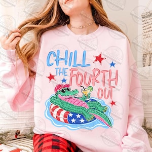 May include: A pale pink sweatshirt with the words "CHILL THE FOURTH OUT" in blue and red. The graphic shows a cartoon alligator wearing sunglasses and a hat, relaxing on a patriotic float with a drink.