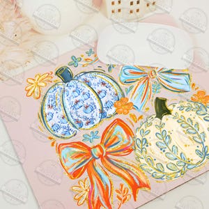May include: A pink surface with watercolor illustrations of pumpkins and bows. The pumpkins are decorated with blue and white patterns, and the bows are orange and blue. Floral accents complete the fall-themed design.