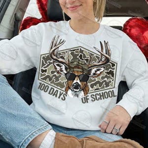 Puede incluir: Sudadera blanca con un gr&aacute;fico de ciervo con gafas de sol y gorra de camuflaje. El gr&aacute;fico est&aacute; dentro de un hex&aacute;gono con estampado de camuflaje, con el texto "100 DAYS OF SCHOOL" debajo. La sudadera es de estilo casual.