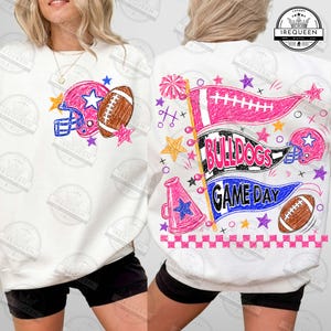 Puede incluir: Una sudadera blanca con un gráfico frontal de un casco de fútbol americano azul y un balón marrón. El gráfico trasero muestra tres banderines con el texto BULLDOGS GAME DAY, un megáfono rosa y un borde a cuadros de color rosa.