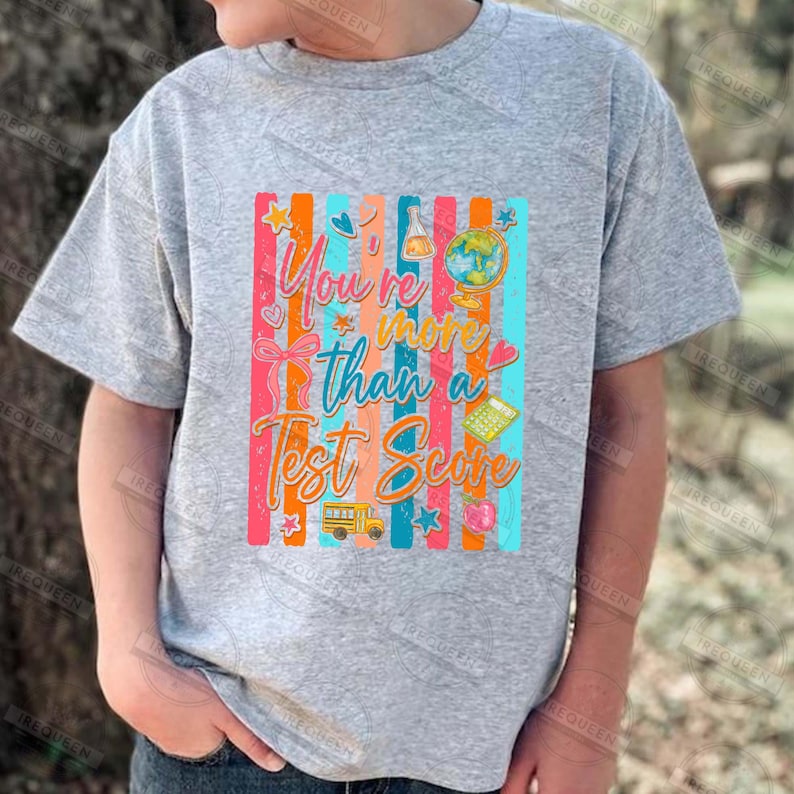 Może przedstawiać: Szara koszulka z kr&oacute;tkim rękawem z kolorową grafiką z napisem "You're more than a Test Score". Projekt zawiera szkolny autobus, globus, kalkulator i inne ikony szkolne na paskowanym tle.