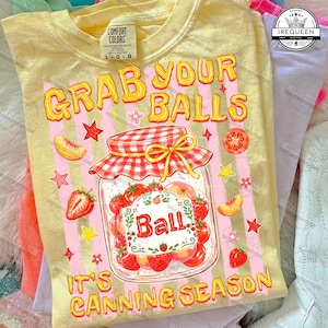 May include: Yellow Comfort Colors t-shirt with the text "GRAB YOUR BALLS" in yellow and red bubble letters. A jar of fruit with the word "Ball" is in the center. The phrase "IT'S CANNING SEASON" is at the bottom.