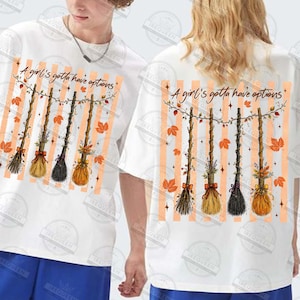 May include: A white t-shirt featuring a graphic print of four hanging broomsticks decorated with autumn leaves and flowers. Above the broomsticks, the text reads A girl's gotta have options. The background of the graphic consists of vertical orange stripes.