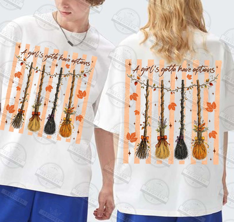 May include: A white t-shirt featuring a graphic print of four hanging broomsticks decorated with autumn leaves and flowers. Above the broomsticks, the text reads A girl's gotta have options. The background of the graphic consists of vertical orange stripes.