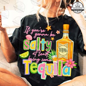 Puede incluir: Camiseta negra con la frase "If you're gonna be salty at least bring some Tequila." El diseño incluye una botella de tequila, un chupito, una rodaja de lima y flores. Prenda de vestir informal.