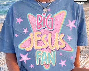May include: A light blue t-shirt featuring a large pink star graphic with the text BIG JESUS FAN in layered, glittery-style lettering. The design is surrounded by smaller pink stars, and the shirt is worn with a beaded necklace featuring a butterfly pendant.