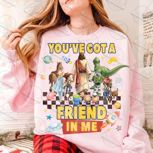 May include: Pink sweatshirt with the text "YOU'VE GOT A FRIEND IN ME" in yellow and red. The design features cartoon characters including Jesus, a dinosaur, and characters from the Toy Story franchise.