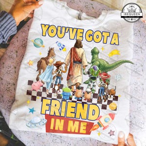 May include: White t-shirt with the text "YOU'VE GOT A FRIEND IN ME" in yellow and red. The design features cartoon characters including Jesus, a dinosaur, and characters from Toy Story. A rocket and a UFO are also included in the design.