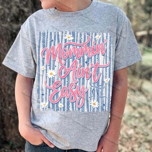 May include: A gray t-shirt featuring the phrase "Mommin' Ain't Easy" in pink script. The design includes a blue and white striped background, white daisies, and a bow graphic. The shirt is short-sleeved.