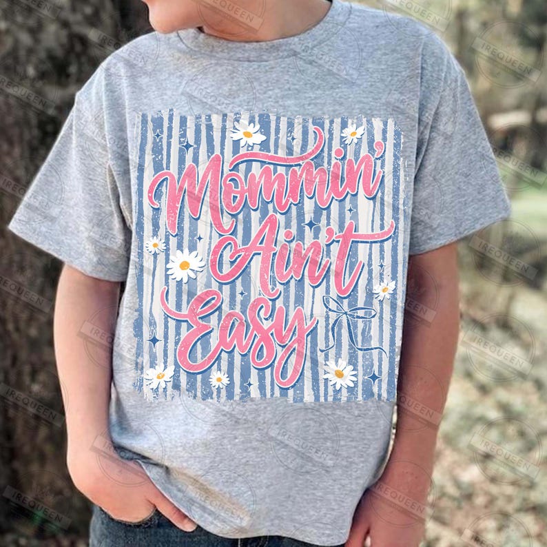 May include: A gray t-shirt featuring the phrase "Mommin' Ain't Easy" in pink script. The design includes a blue and white striped background, white daisies, and a bow graphic. The shirt is short-sleeved.