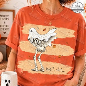 May include: An orange t-shirt featuring a graphic of a skeleton wearing a white ghost sheet, bent over with hands on its hips. The design includes scattered autumn leaves and the text "Well, shit..." printed in a black script font at the bottom.
